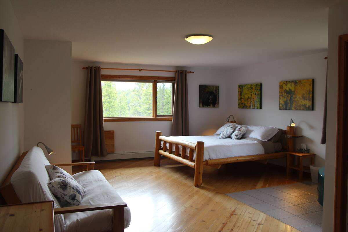 A bedroom with a wooden bed, a wooden chair, a wooden desk, a window with curtains, and two paintings on the wall.