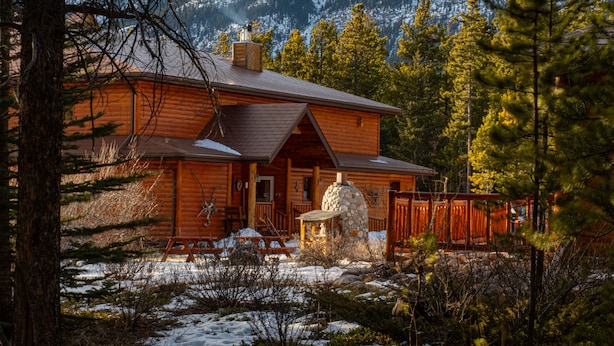 A log cabin with a stone fireplace, surrounded by snow and trees, with mountains in the background. - Aurum Lodge (Cline River)