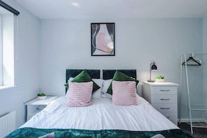 Une chambre à coucher comprenant un lit, des oreillers, une table de chevet, une lampe, un radiateur et une œuvre d’art accrochée au mur.