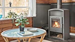 A cozy room with a wood-burning stove, a round glass table with a book and a plant, and a window with a view of trees. - Beautiful home in Frederiksværk (Frederiksværk)
