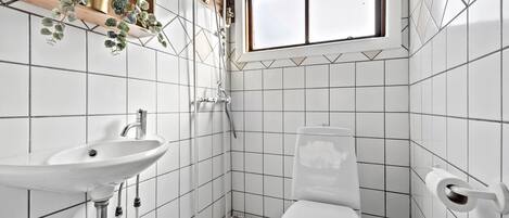 A small, tiled bathroom with a white sink, toilet, and shower. There is a window above the toilet and a plant on the wall.