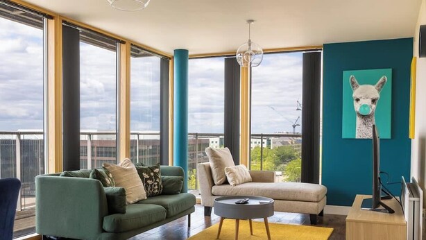 Un salon moderne avec de grandes fenêtres, un canapé vert, un canapé beige et un tableau représentant un lama.