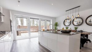 A modern kitchen with white cabinets, a large island, and a view of the outdoors through sliding glass doors. - 5-Bed Home | Canal Views | Parking for 2+ Cars (Milton Keynes)