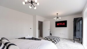 A modern hotel room with a large bed, a side table, a chair, a TV mounted on the wall, and a door leading to another room. - 5-Bed Home | Canal Views | Parking for 2+ Cars (Milton Keynes)