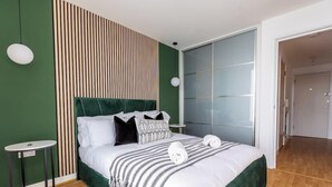 Ett modernt hotellrum med en accentvägg i grönt, en stor säng med vita sängkläder, en randig pläd och utsikt över en stadssiluett genom ett stort fönster.