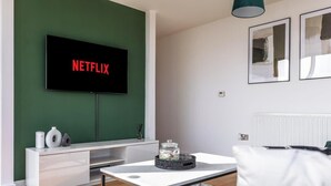Ett modernt vardagsrum med en platt-TV som visar "NETFLIX", ett vitt konsolbord med dekorativa föremål, en vit soffa och en grön accentvägg.
