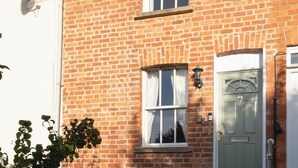 Un bâtiment en briques avec une porte verte portant le numéro 7, une fenêtre avec des rideaux blancs et une petite plante en pot placée à gauche.