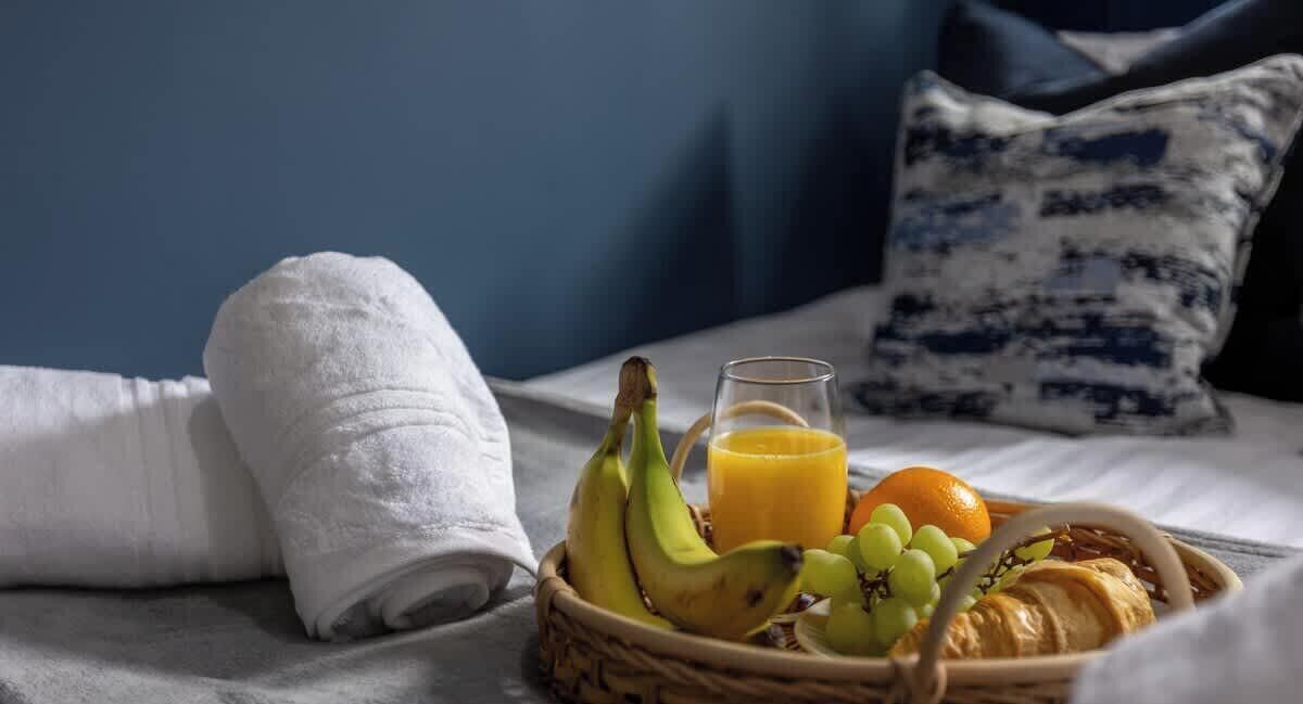 A neatly made bed with a white towel, a basket of breakfast items including bananas, grapes, an orange, and a croissant, and a glass of orange juice.