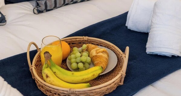 A wicker basket with bananas, grapes, an orange, and a croissant on a bed with a blue towel and white linens. - Central MK Apartment with Gated Parking & Fast Wi-Fi (Milton Keynes)