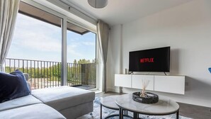 Een moderne woonkamer met een bank, een salontafel, een aan een witte kast bevestigde televisie, en een balkon met uitzicht op groen.