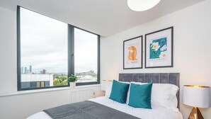 Ein modernes Schlafzimmer mit großem Fenster, einem Bett mit türkisfarbenen Kissen und zwei gerahmten Kunstwerken an der Wand.