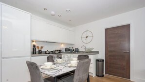 Eine kompakte Küche mit weißen Schränken, einer Mikrowelle und einem Spülbecken. Ein Esstisch ist gedeckt mit Tellern, Gläsern und Besteck. Eine Uhr ist an der Wand befestigt, und ein Mülleimer ist sichtbar.