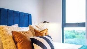 A bed with a blue headboard, multiple pillows, and a view of greenery through a window. - Modern, Spotless & Close to City Hotspots! (Greater Manchester)
