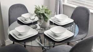 A round glass table with white square plates and clear wine glasses, surrounded by grey chairs. - Spacious 2-Bedroom Home with Parking & Wi-Fi (Milton Keynes)