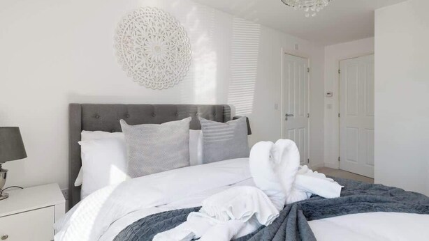 A neatly made bed with white and gray bedding, a gray headboard, and a decorative wall piece. - Spacious 2-Bedroom Home with Parking & Wi-Fi (Milton Keynes)
