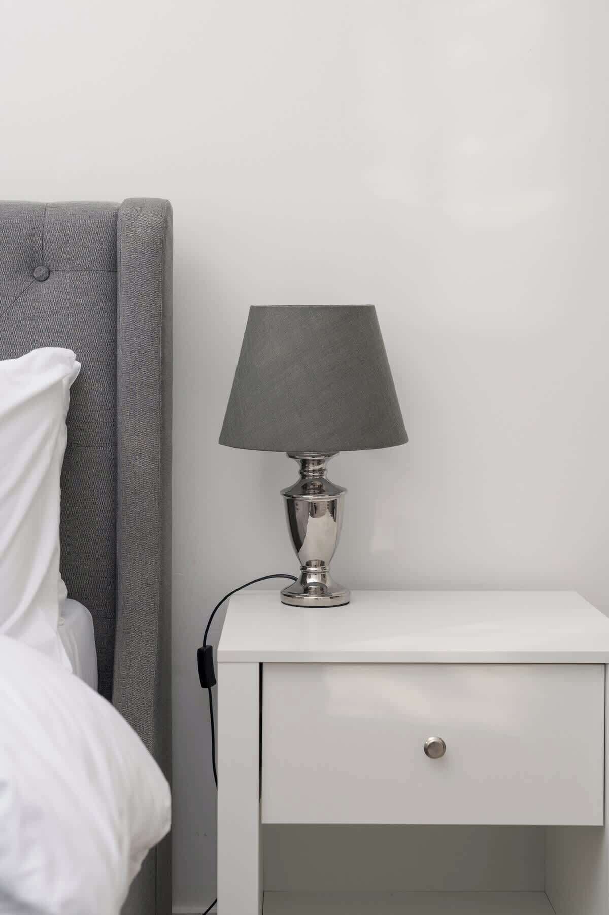 Une table de chevet blanche avec une lampe, une tête de lit grise et un oreiller blanc.