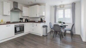 A modern kitchen with white cabinets, a stainless steel range hood, and a dining area with a table and chairs. - Spacious 2-Bedroom Home with Parking & Wi-Fi (Milton Keynes)