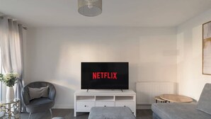 A modern living room with a flat-screen TV displaying "NETFLIX," a grey sofa, a white entertainment unit, and a grey armchair. - Spacious 2-Bedroom Home with Parking & Wi-Fi (Milton Keynes)