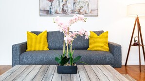 En grå soffa med gula kuddar, en svart, rektangulär blomkruka med gröna växter och ett träbord.