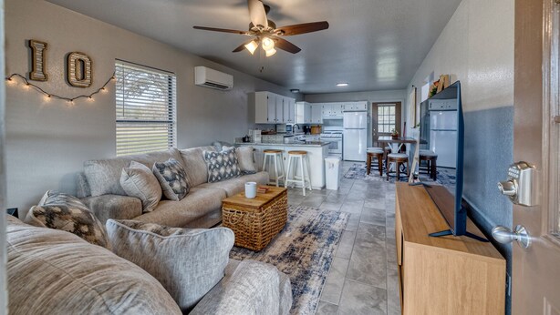 A spacious living area with a sofa, a coffee table, and a TV stand. There is a kitchen area with a refrigerator, stove, and bar stools. - Live Oak Cottage on a beautiful lot with large oak trees and fenced backyard. (Concan)