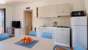 A compact kitchen with a microwave, stove, sink, and cabinets. There is a dining table with chairs and a bowl of fruit.