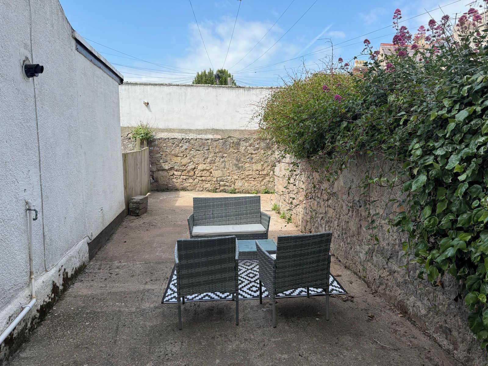 A patio with two wicker chairs and a table, a stone wall, and a white building.