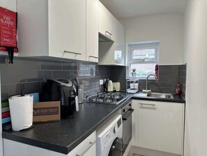 Une cuisine moderne avec des meubles blancs, un plan de travail noir, une cuisinière à gaz et une fenêtre équipée de stores.