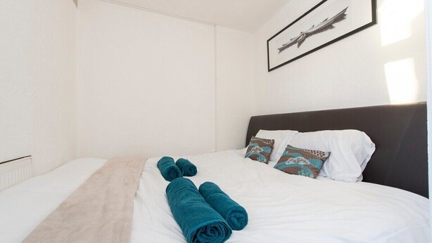 A neatly made bed with white linens and teal towels, a dark headboard, and a framed picture on the wall. - Cosy haven in Harwich (Essex)