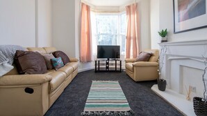 A living room with a beige sofa, a television, a fireplace, and a window with curtains. - Cosy haven in Harwich (Essex)