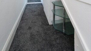 A narrow hallway with a carpeted floor leading to a bedroom with a bed and a small shelf. - Cosy haven in Harwich (Essex)