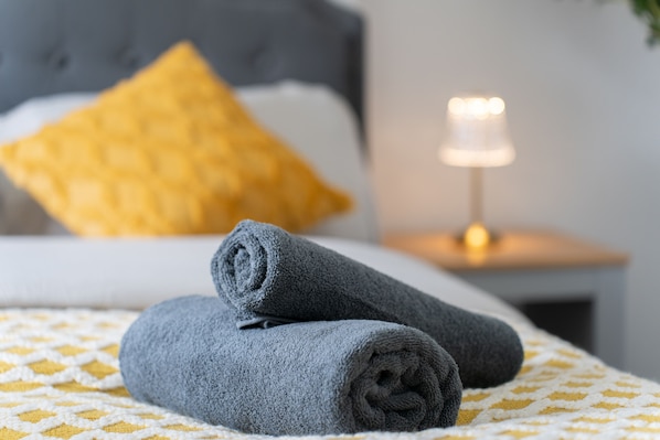 Un lit bien fait, avec une serviette grise roulée, un coussin jaune à motifs et une table de chevet sur laquelle brûle une bougie.