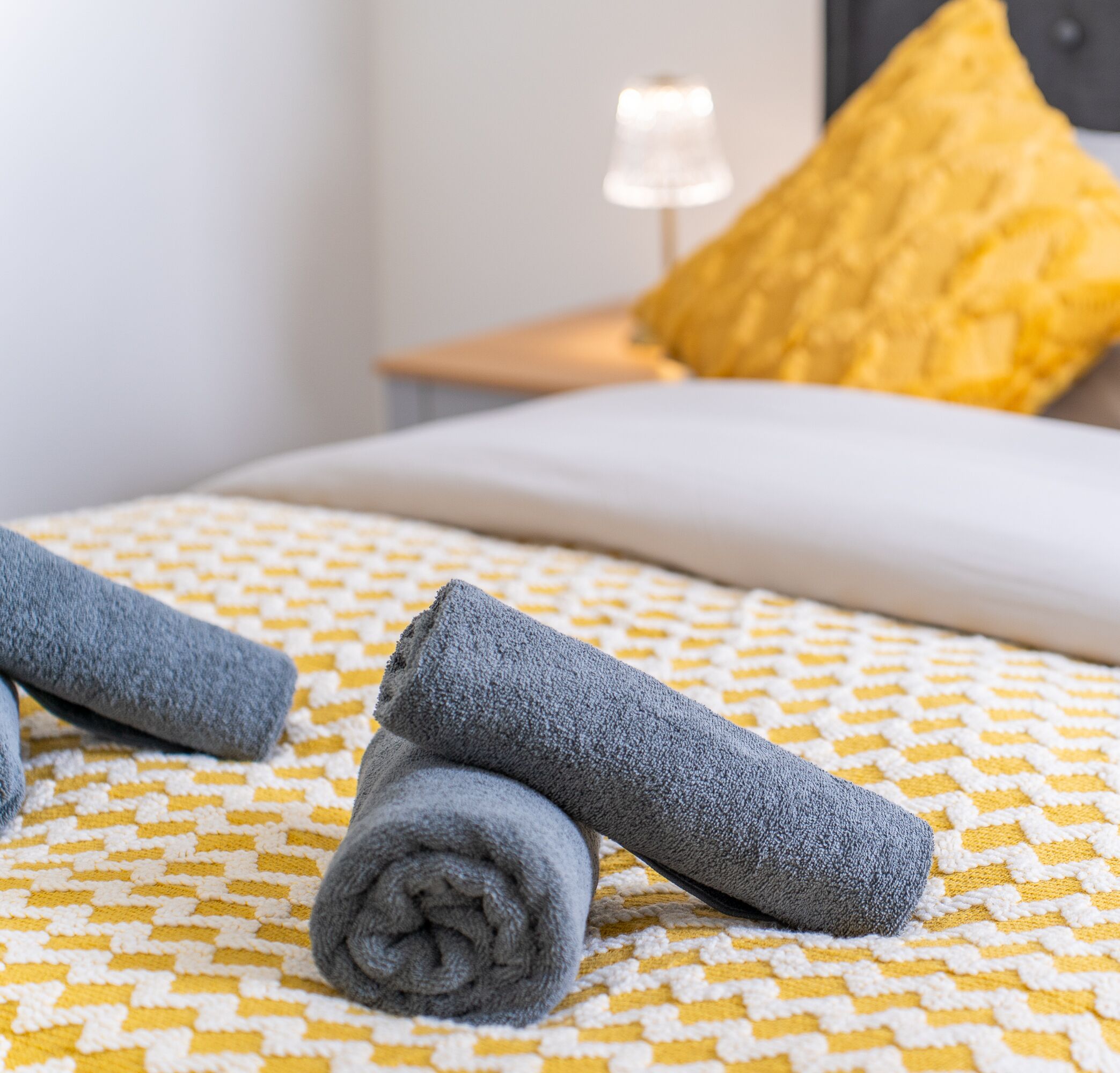 Un lit bien fait, avec une couvre-lit à motifs, deux serviettes enroulées, un oreiller jaune et une lampe de chevet.