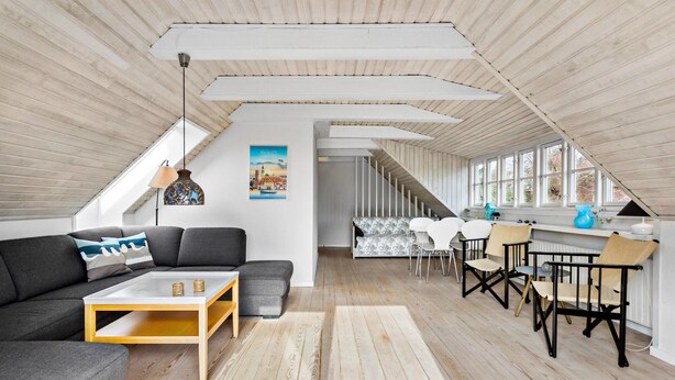 Ein geräumiges Dachzimmer mit hoher Decke, Holzbalken, einer Couch, einem Esstisch mit Stühlen und Blick durch große Fenster.