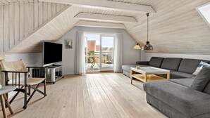 Ein geräumiges Loft mit hohen Decken, Holzbalken, einem großen Fernseher, einem grauen Sofa, einem kleinen Tisch und Blick auf die Stadt durch eine Schiebeglastür.