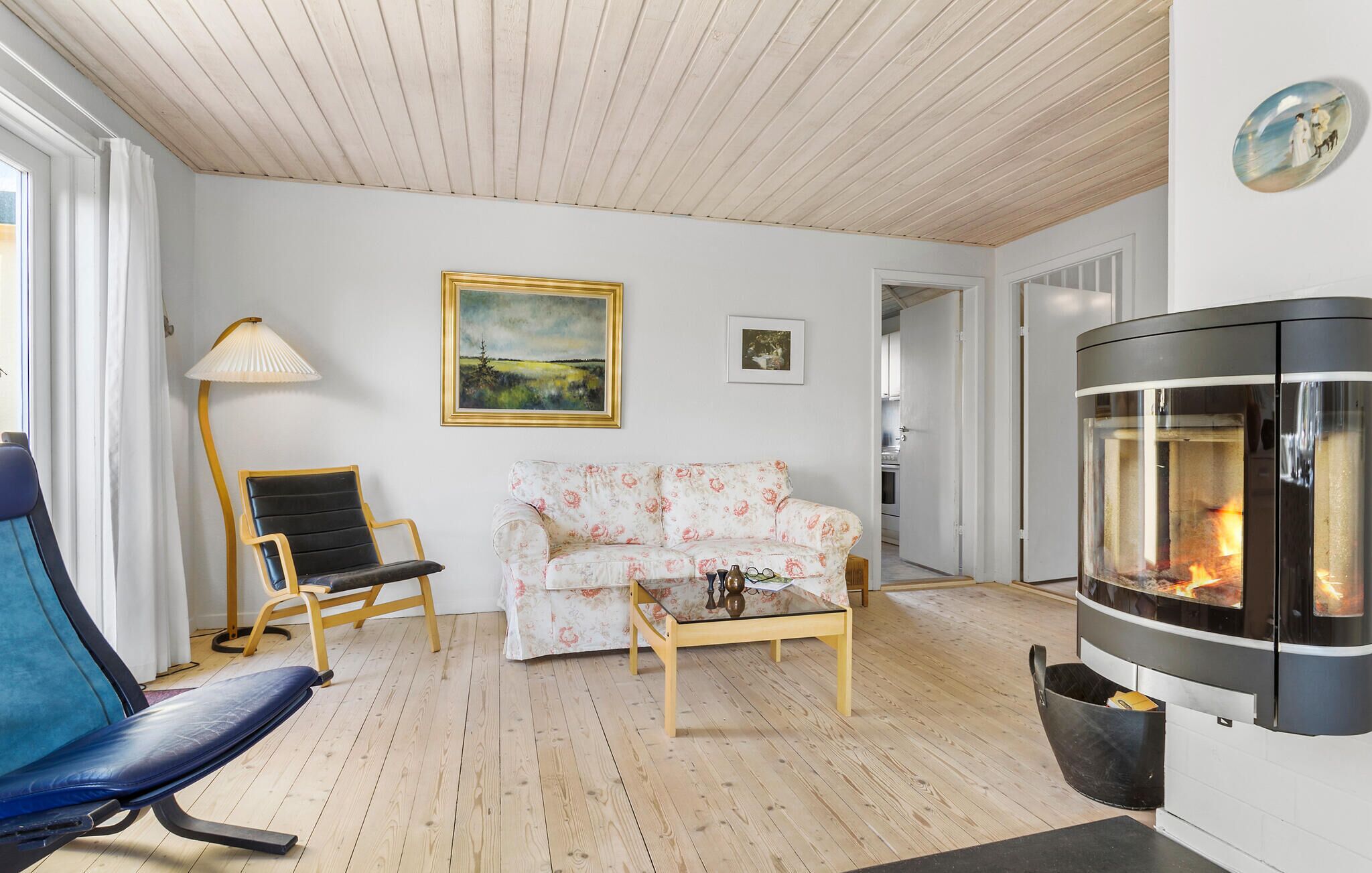Ett vardagsrum med en soffa, en fåtölj, ett soffbord i glas, en öppen spis och en tavla på väggen.