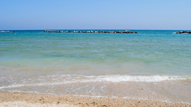 A beach with clear turquoise water, pebbly shore, and a rocky breakwater in the distance.