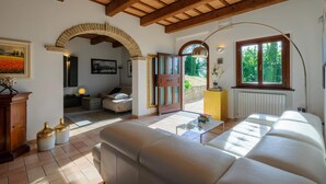 A spacious living room with a large sofa, a coffee table, and a fireplace. There is a wooden ceiling and a stone archway leading to another room.