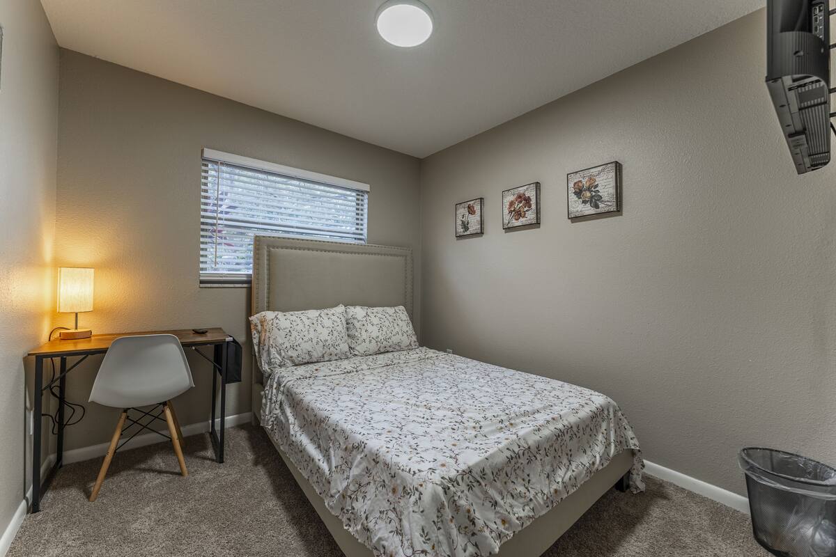 A bedroom with a bed, a desk, a chair, a window with blinds, and a wall-mounted heater.