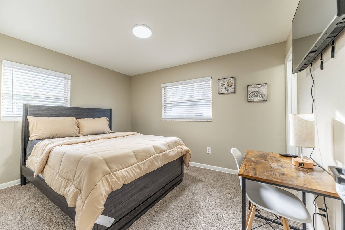 A bedroom with a bed, a desk, and a chair. There are two windows with blinds, a ceiling light, and a wall-mounted air conditioner.