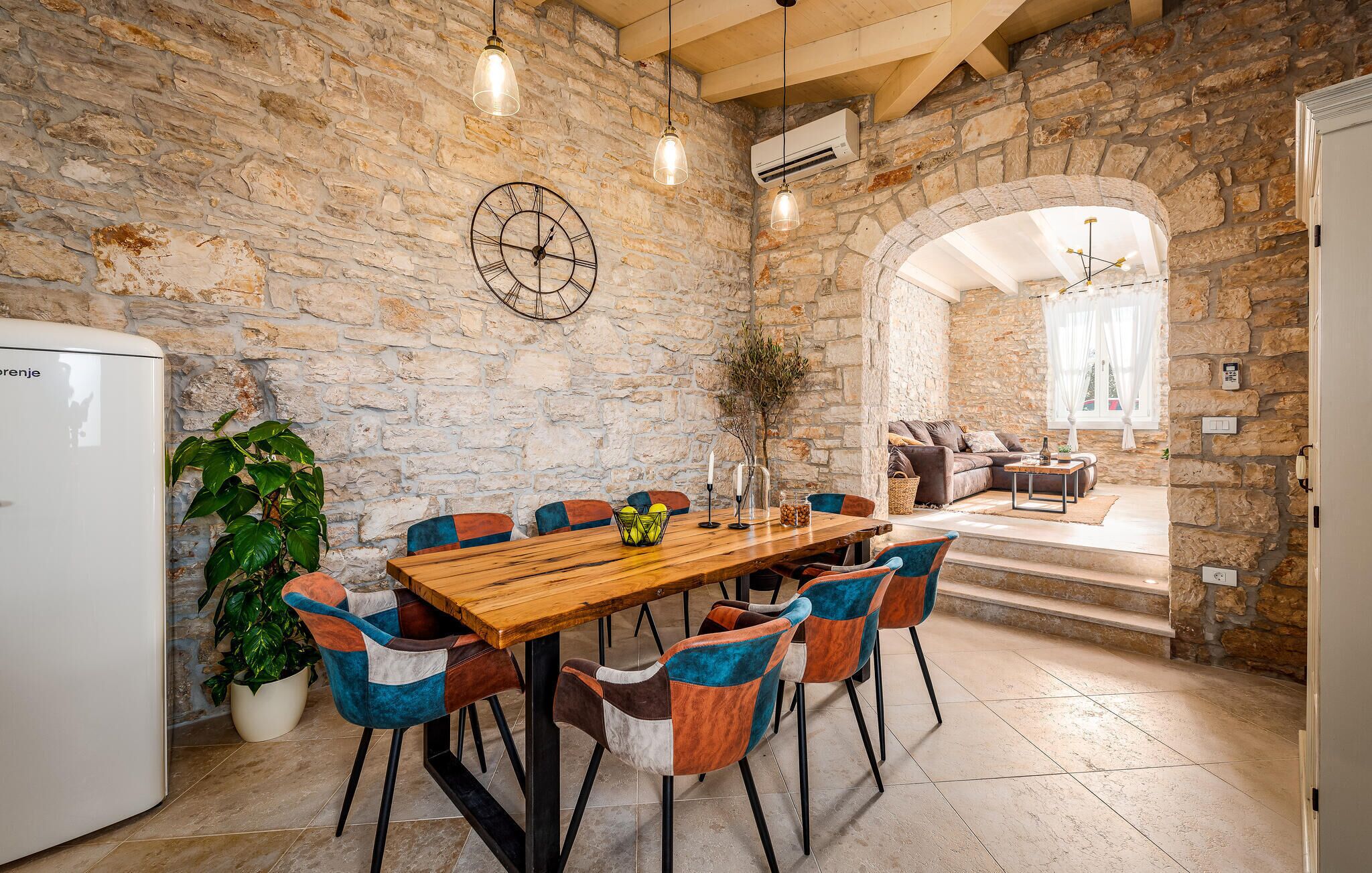 A rustic stone-walled room with a wooden dining table, colorful chairs, a clock, and a cozy seating area with a sofa and cushions.
