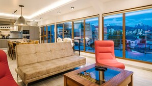 A modern living room with a sofa, a red chair, and a coffee table. There is a kitchen area with a dining table and chairs. Large windows offer a view of a mountainous landscape. - Gente del Sur - Fique (Ushuaia)