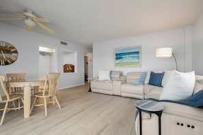A living room with a beige sofa, a dining table with chairs, a clock, and a painting of a beach scene. - Oceanfront Oasis - 2BR/2BA direct oceanfront! (Satellite Beach)