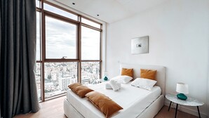 A modern hotel room with a large window, a bed with white linens, two pillows, a small bedside table with a lamp, and a city view. - WealthyStays: SkyHigh Central (Nicosia)