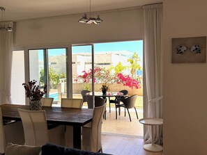 A dining area with a table and chairs, a vase with dried flowers, and a view of a pool and buildings outside.