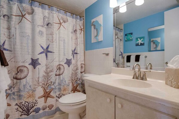 A bathroom with a nautical theme, featuring a shower curtain with sea creatures, a toilet, a sink with a mirror, and a towel rack.