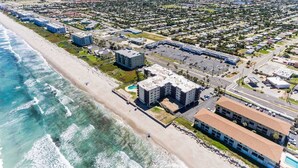 An aerial view of a coastal area with a beach, a parking lot, and several buildings including a hotel with a pool. - You cannot get any closer to the beach! 2BR condo (Satellite Beach)