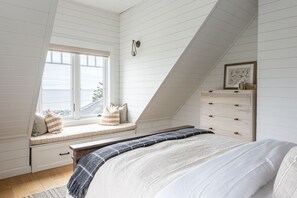 Un dormitorio con techo inclinado, una cama con manta blanca y gris, un banco con almohadas junto a una ventana y un aparador con cajones.