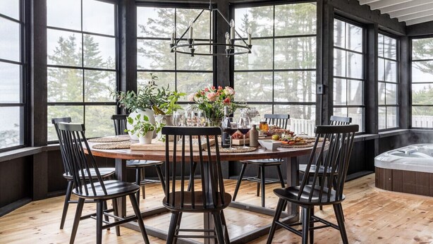 Une table de salle à manger ronde, entourée de six chaises, et de grandes fenêtres qui donnent sur un paysage arboré.