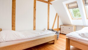 Ein Schlafzimmer mit zwei Holzbettgestellen übereinander, einer kleinen weißen Kommode und einem Fenster mit Blick auf Grünflächen.