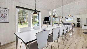 A spacious dining area with a long white table and modern chairs, large windows offering a view of the outdoors, and pendant lighting.